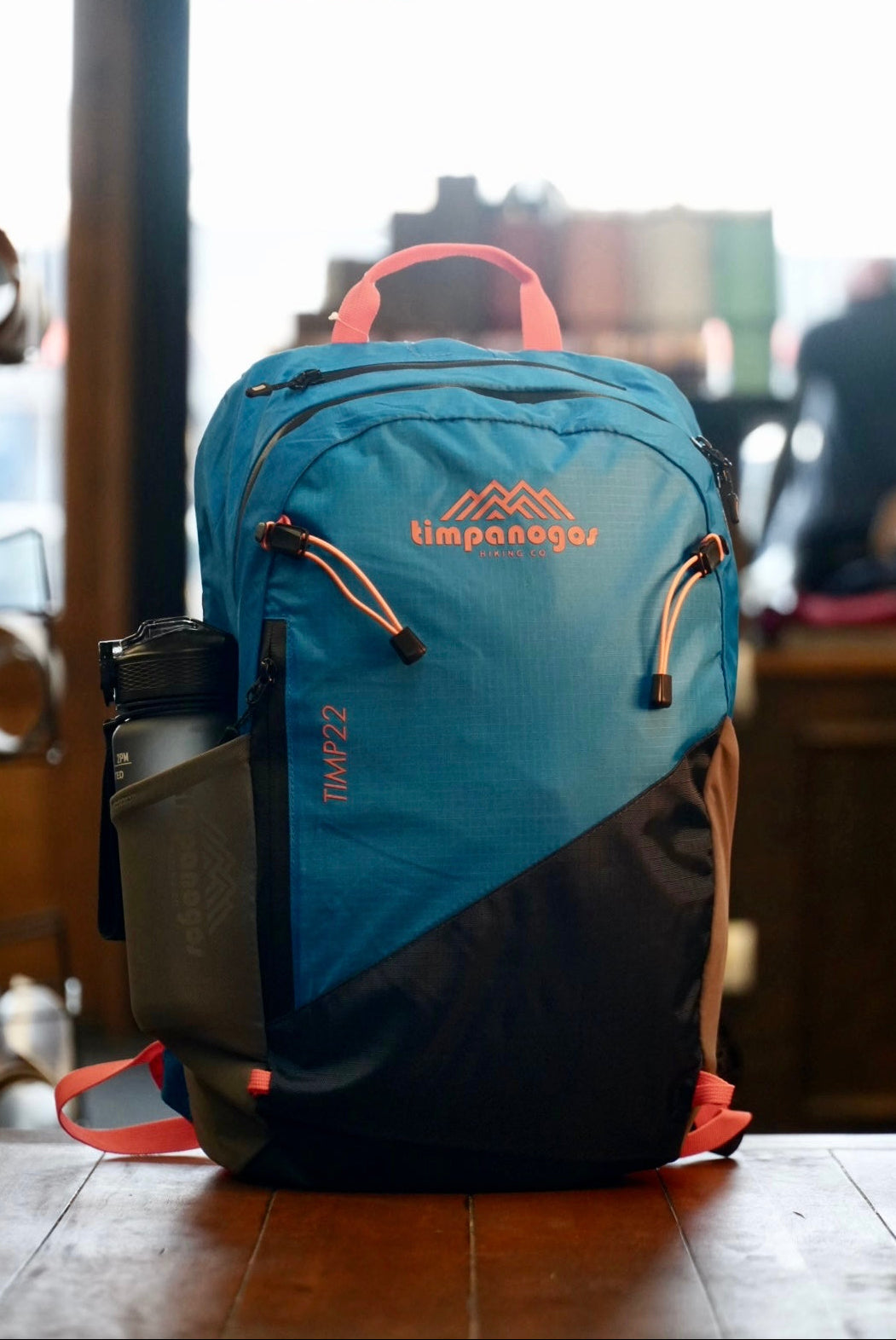 Blue Timpanogos backpack with coral accents on a wooden surface, blurred indoor background