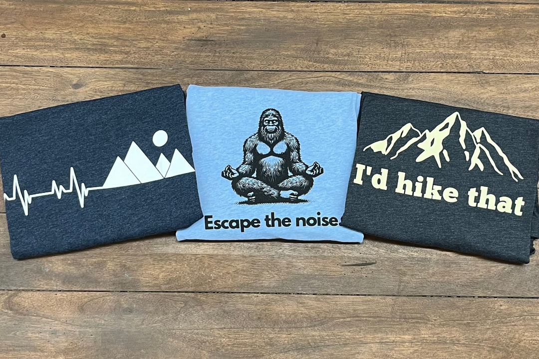 Three shirts with mountain and nature-themed designs on a wooden surface
