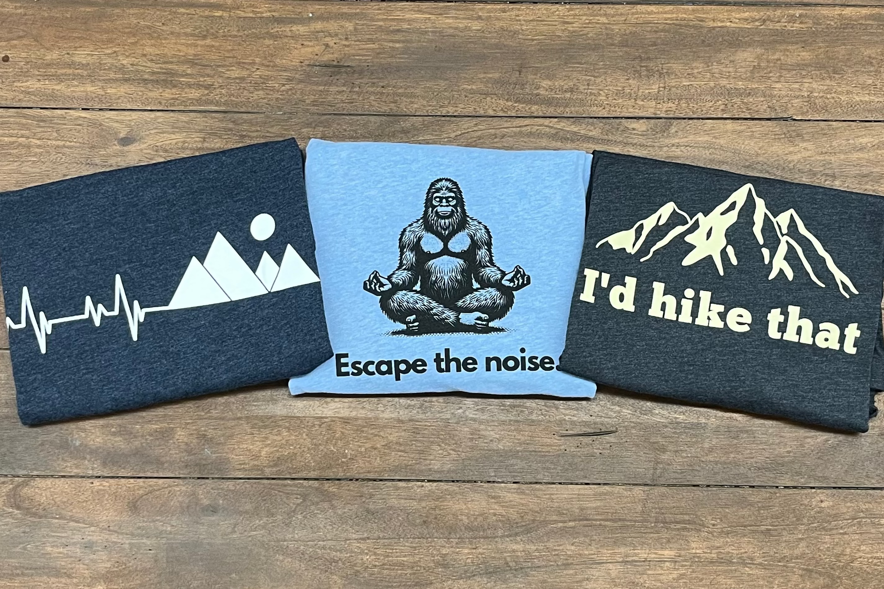 Three shirts with mountain and nature-themed designs on a wooden surface