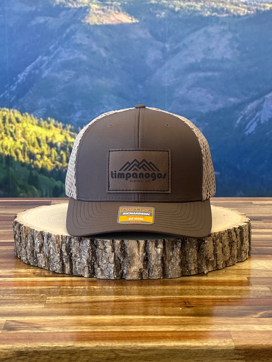 Heather Brown trucker hat with a beige mesh back, featuring a brown leather patch with a mountain graphic and "Timpanogos Hiking Co." text, displayed on a rustic wood surface with a mountain backdrop