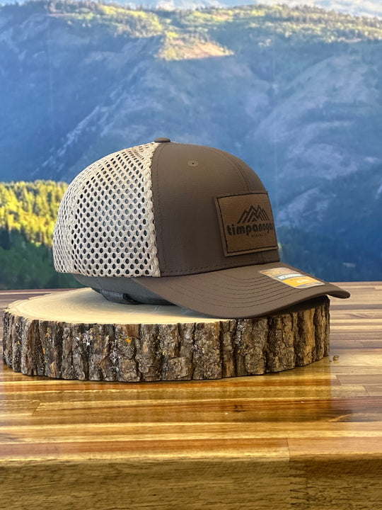 Heather Brown trucker hat with a beige mesh back, featuring a brown leather patch with a mountain graphic and "Timpanogos Hiking Co." text, displayed on a rustic wood surface with a mountain backdrop