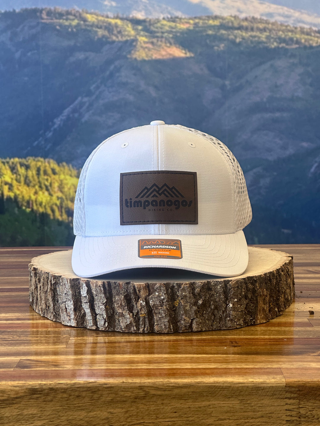 White trucker cap with a brown leather patch showcasing a mountain design and "Timpanogos Hiking Co." text, placed on a wood slab with a scenic mountain view behind it
