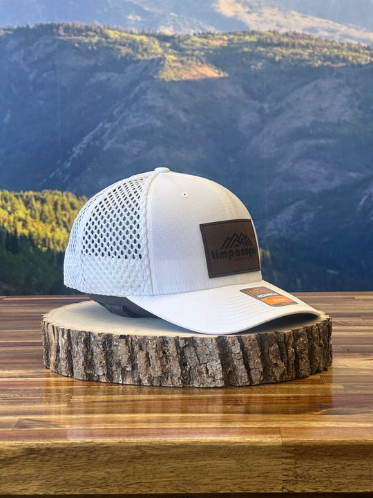 White trucker cap with a brown leather patch showcasing a mountain design and "Timpanogos Hiking Co." text, placed on a wood slab with a scenic mountain view behind it