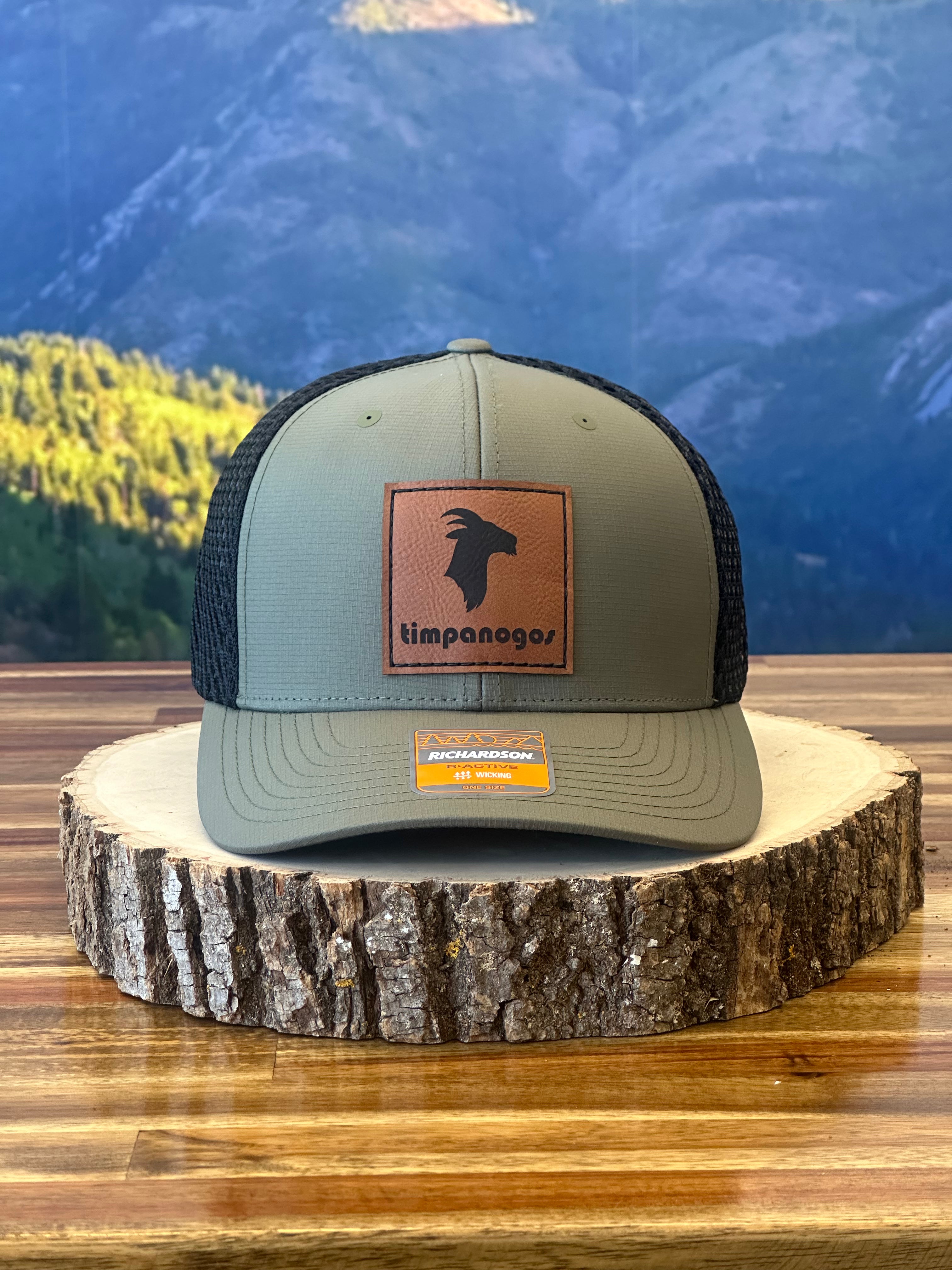 Heather Military Green trucker hat with a brown leather Timpanogos Hiking Co. goat logo patch, displayed on a wooden stump with the Wasatch Mountains in the background.