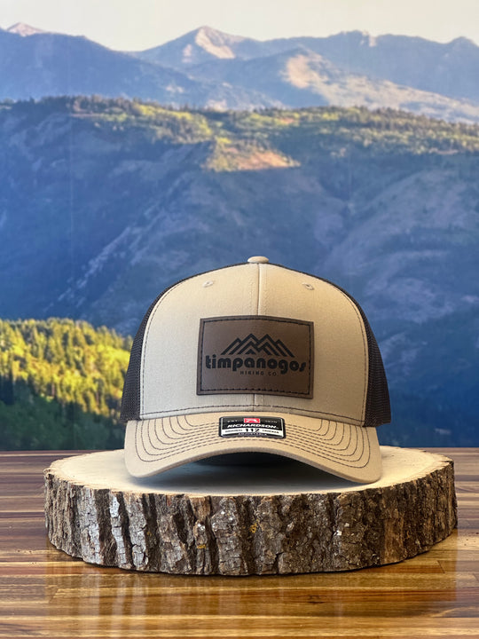 Heather Cream trucker hat with a brown mesh back featuring a brown leather patch with a mountain graphic and "Timpanogos Hiking Co." text, displayed on a rustic wood surface with a mountain backdrop