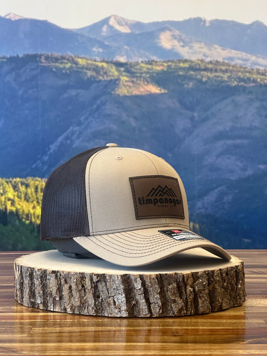 Heather Tan trucker hat with a brown mesh back, featuring a brown leather patch with a mountain graphic and "Timpanogos Hiking Co." text, displayed on a rustic wood surface with a mountain backdrop