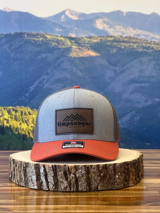 Heather Gray and orange trucker hat featuring a brown leather patch with a mountain graphic and "Timpanogos Hiking Co." text, displayed on a rustic wood surface with a mountain backdrop