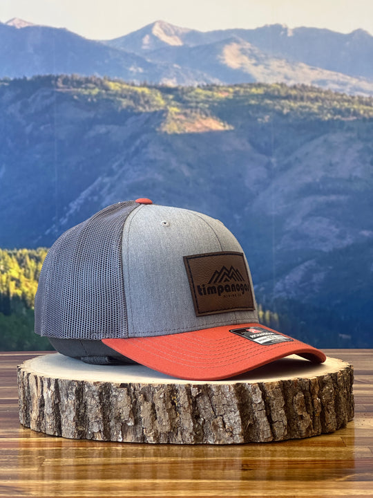 Heather Gray trucker hat with an orange brim and a brown leather patch featuring a mountain graphic and "Timpanogos Hiking Co." text, displayed on a rustic wood surface with a mountain backdrop