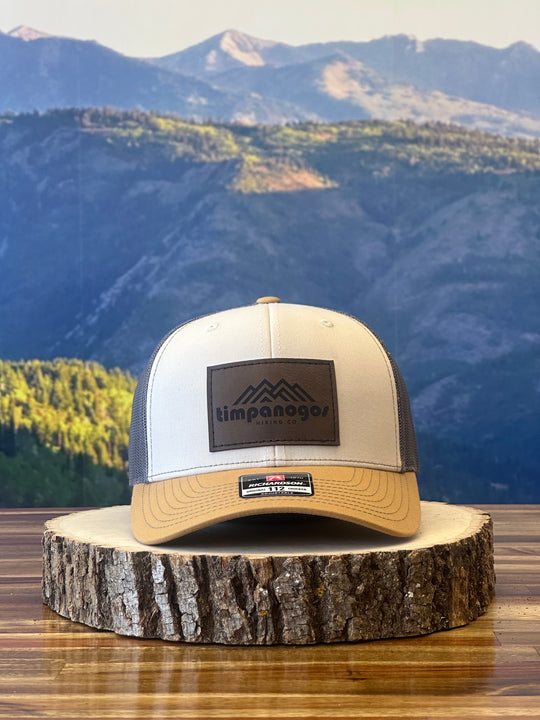 Heather Cream and tan trucker hat featuring a brown leather patch with a mountain graphic and "Timpanogos Hiking Co." text, displayed on a rustic wood surface with a mountain backdrop