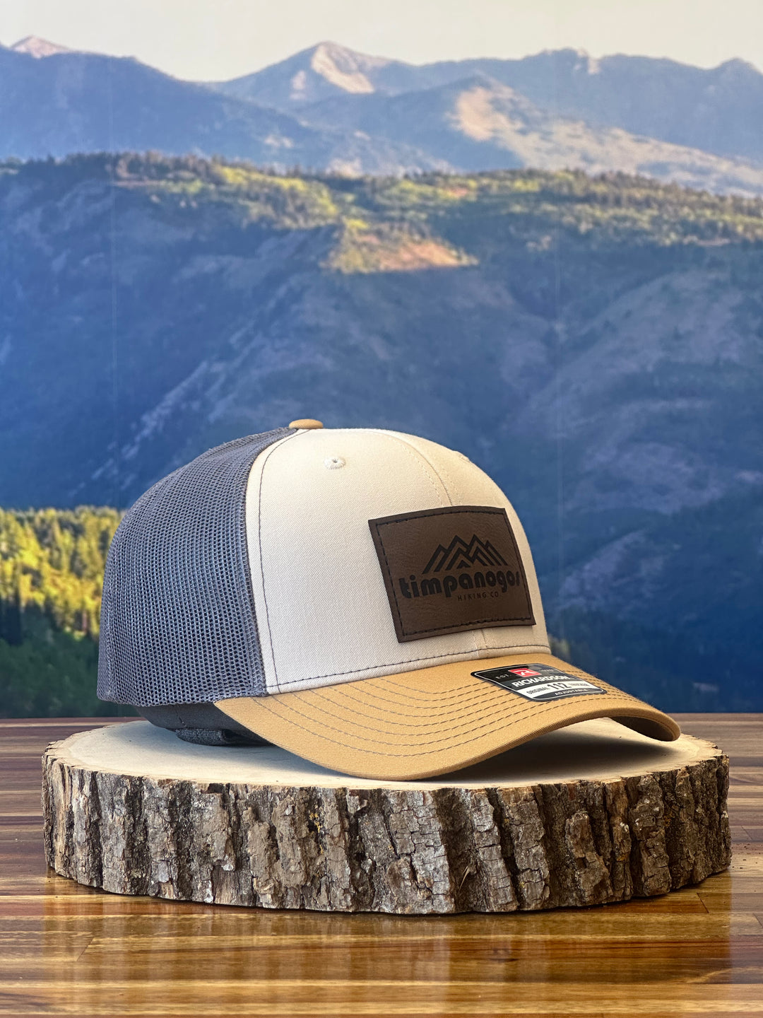 Heather Cream and tan trucker hat with a brown leather patch featuring a mountain graphic and "Timpanogos Hiking Co." text, displayed on a rustic wood surface with a mountain backdrop