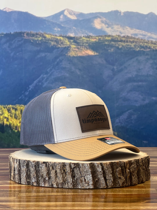 Heather Cream and tan trucker hat with a brown leather patch featuring a mountain graphic and "Timpanogos Hiking Co." text, displayed on a rustic wood surface with a mountain backdrop