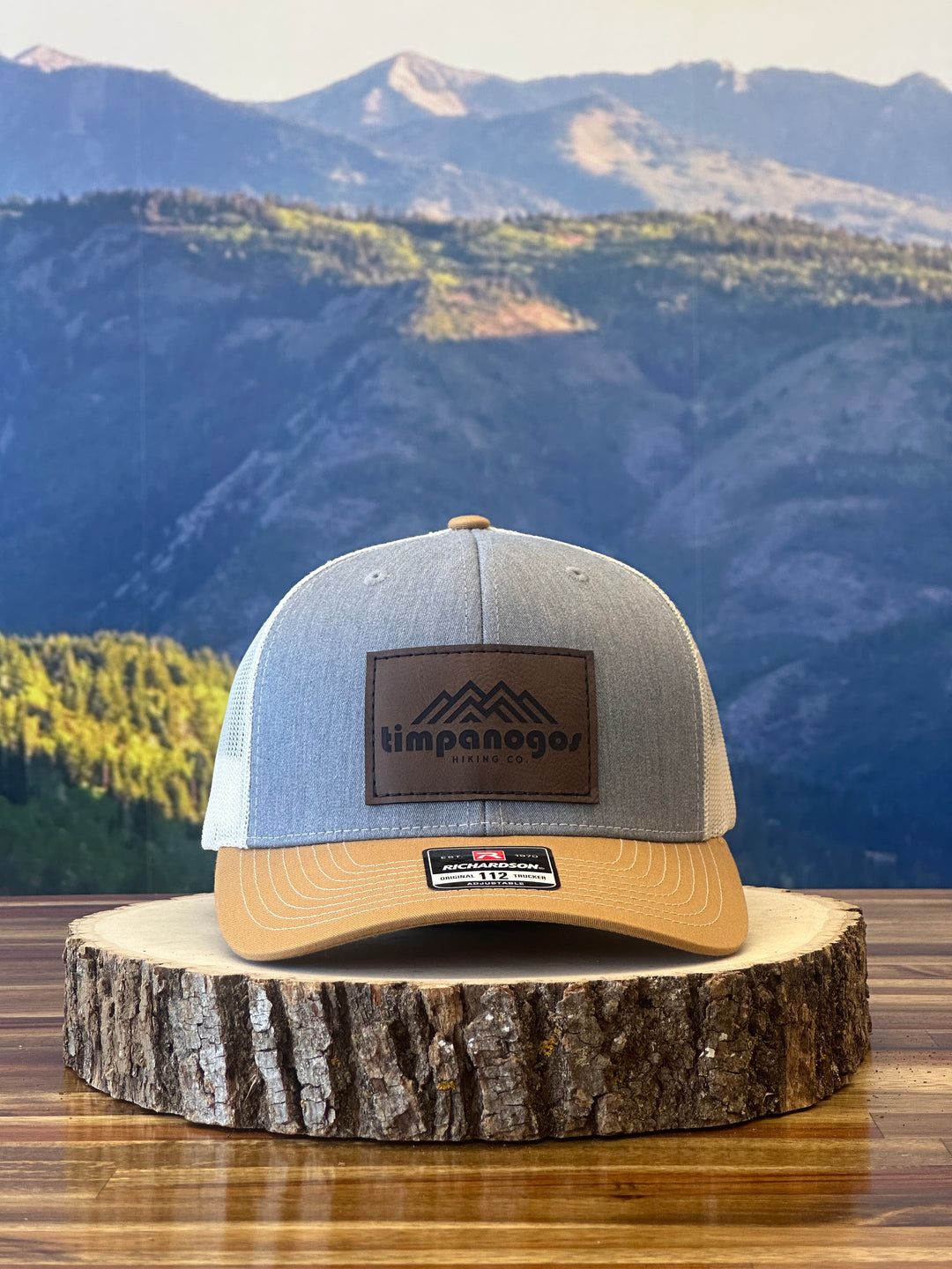 Heather Gray and tan trucker hat featuring a brown leather patch with a mountain graphic and "Timpanogos Hiking Co." text, displayed on a rustic wood surface with a mountain backdrop