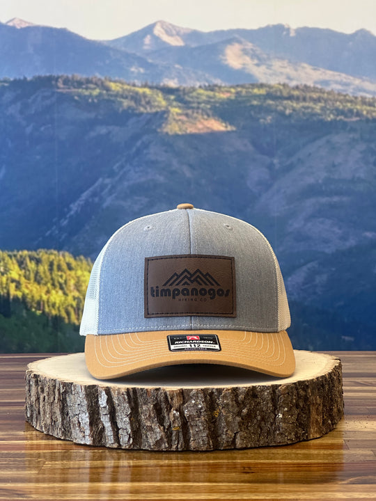 Heather Gray and tan trucker hat featuring a brown leather patch with a mountain graphic and "Timpanogos Hiking Co." text, displayed on a rustic wood surface with a mountain backdrop