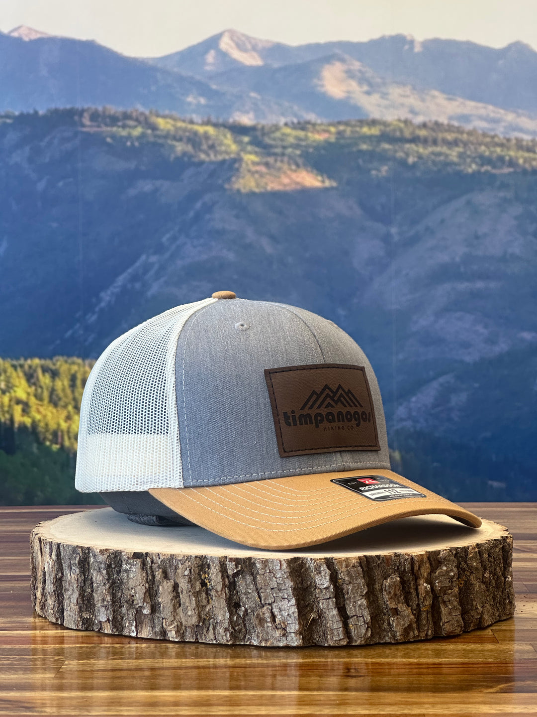 Heather Gray and tan trucker hat featuring a brown leather patch with a mountain graphic and "Timpanogos Hiking Co." text, displayed on a rustic wood surface with a mountain backdrop