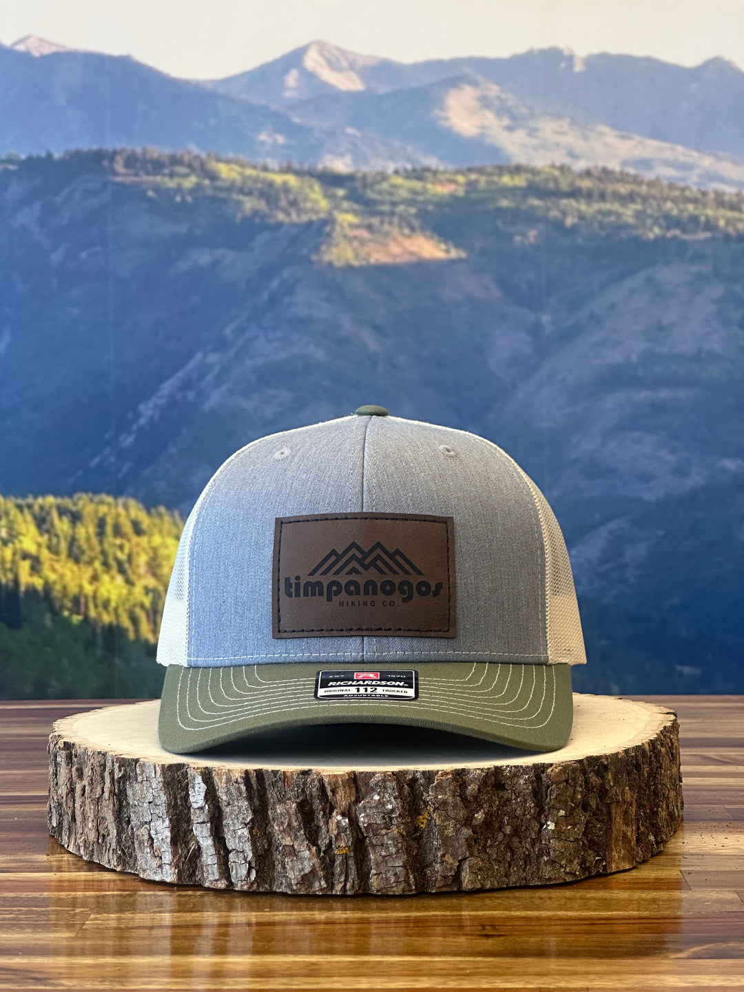 Heather Gray and olive green trucker hat with a brown leather patch featuring a mountain graphic and "Timpanogos Hiking Co." text, displayed on a rustic wood surface with a mountain backdrop