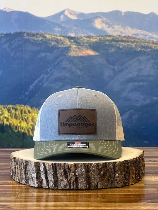 Heather Gray and olive green trucker hat with a brown leather patch featuring a mountain graphic and "Timpanogos Hiking Co." text, displayed on a rustic wood surface with a mountain backdrop