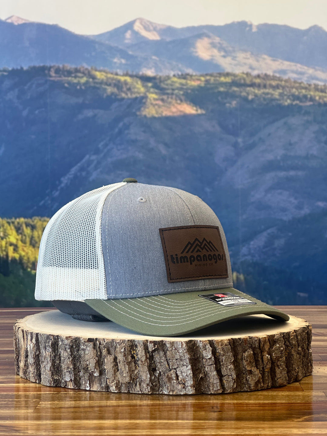 Heather Gray and olive green trucker hat with a brown leather patch featuring a mountain graphic and "Timpanogos Hiking Co." text, displayed on a rustic wood surface with a mountain backdrop