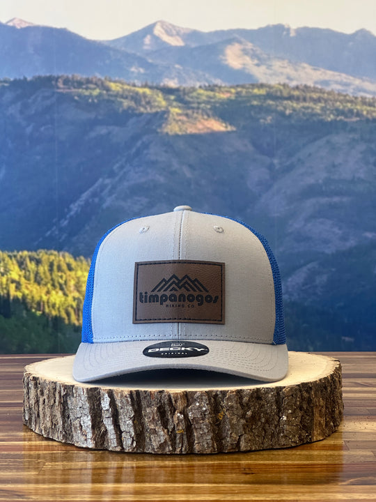 Heather Gray and blue trucker hat featuring a brown leather patch with a mountain graphic and "Timpanogos Hiking Co." text, displayed on a rustic wood surface with a mountain backdrop