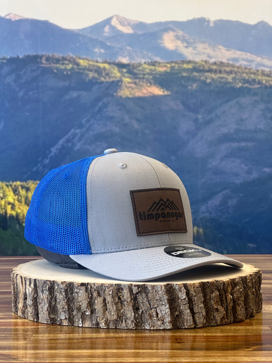 Heather Gray and blue trucker hat featuring a brown leather patch with a mountain graphic and "Timpanogos Hiking Co." text, displayed on a rustic wood surface with a mountain backdrop