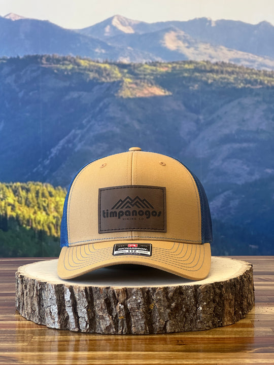 Heather Tan and blue trucker hat featuring a brown leather patch with a mountain graphic and "Timpanogos Hiking Co." text, displayed on a rustic wood surface with a mountain backdrop


