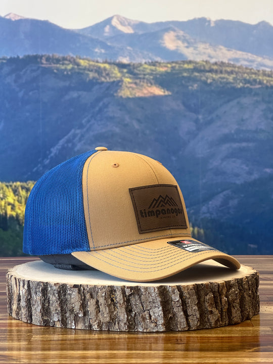 Tan and blue trucker cap with a leather patch showcasing a mountain design and "Timpanogos Hiking Co." branding, set against a wooden platform with a scenic mountain view