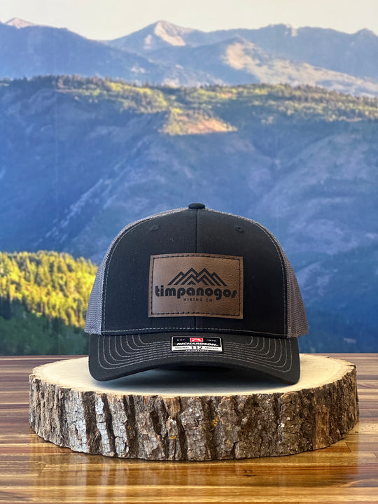 Heather Black trucker hat featuring a brown leather patch with a mountain graphic and "Timpanogos Hiking Co." text, displayed on a rustic wood surface with a mountain backdrop