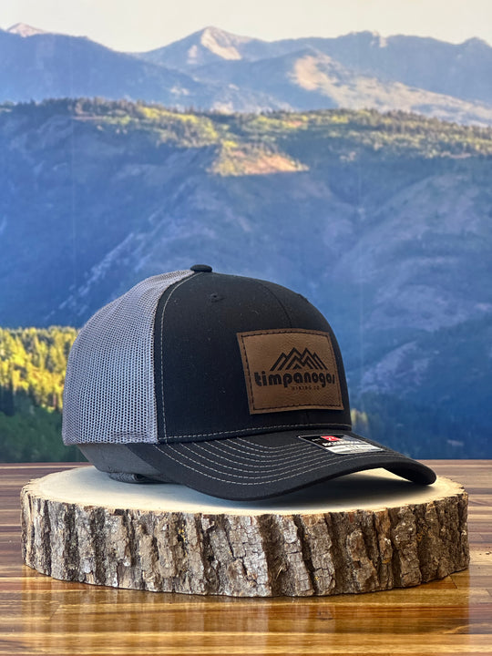Heather Black trucker hat with a gray mesh back, featuring a brown leather patch with a mountain graphic and "Timpanogos Hiking Co." text, displayed on a rustic wood surface with a mountain backdrop