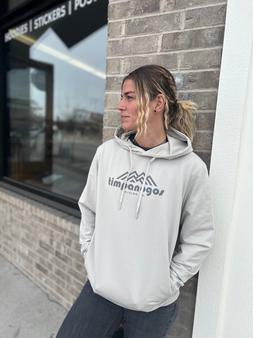 Woman wearing a Heather Cream hoodie with the Timpanogos Hiking Co. mountain logo, standing outdoors against a brick wall with a storefront in the background, showcasing the casual outdoor style of Timpanogos Hiking Co.

