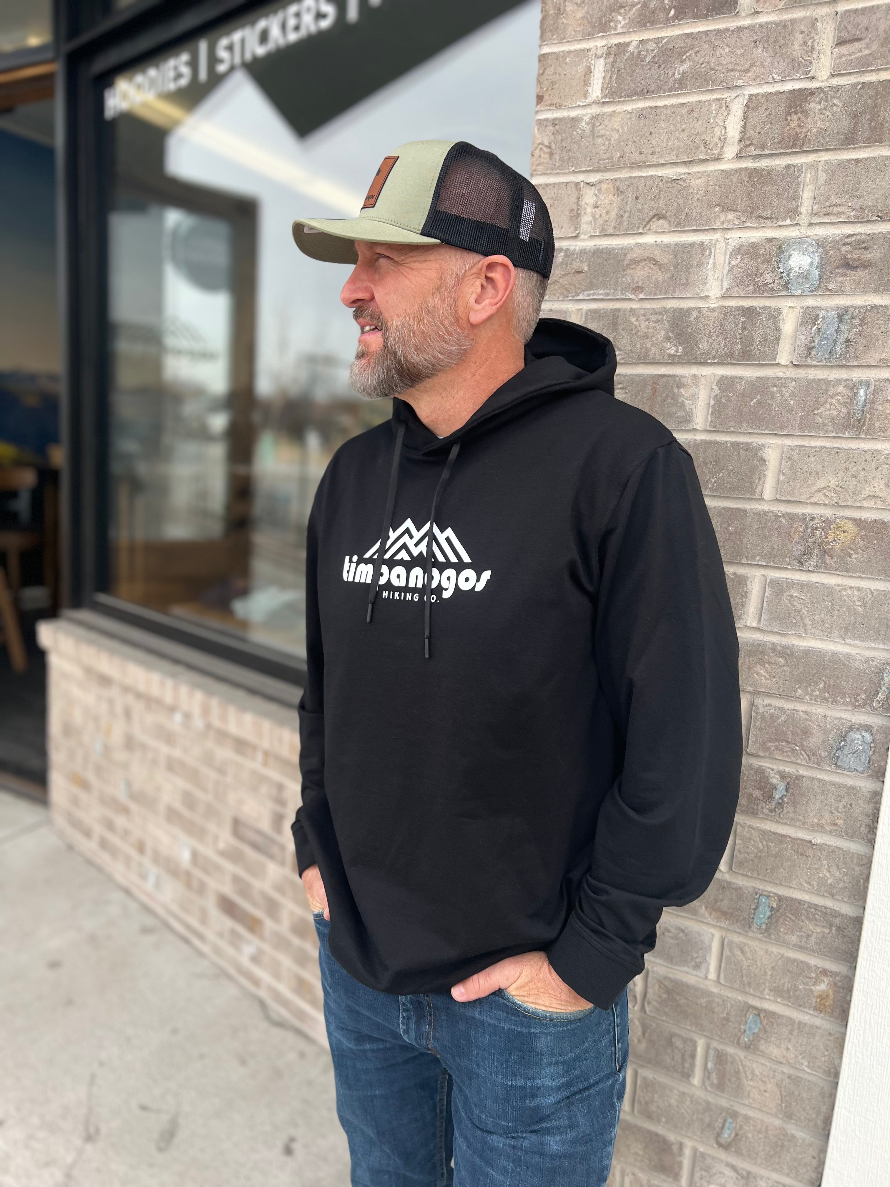 Man wearing a black hoodie with the Timpanogos Hiking Co. logo, paired with a green and black trucker hat, standing outdoors against a brick wall, showcasing the rugged outdoor style of Timpanogos Hiking Co.

