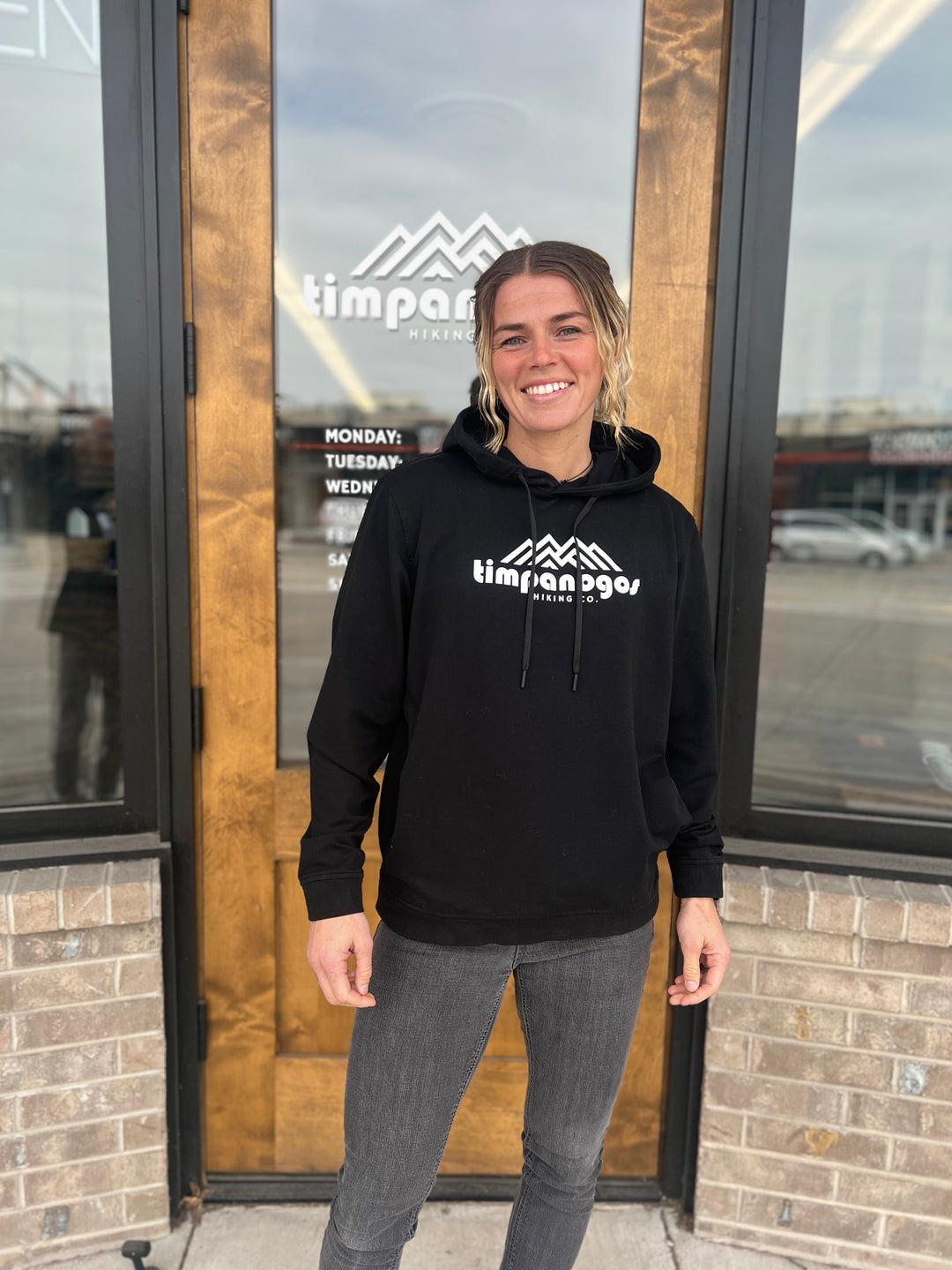 Woman smiling in a black hoodie with the Timpanogos Hiking Co. mountain logo, standing in front of a storefront with a wooden door, showcasing the adventurous outdoor style of Timpanogos Hiking Co.

