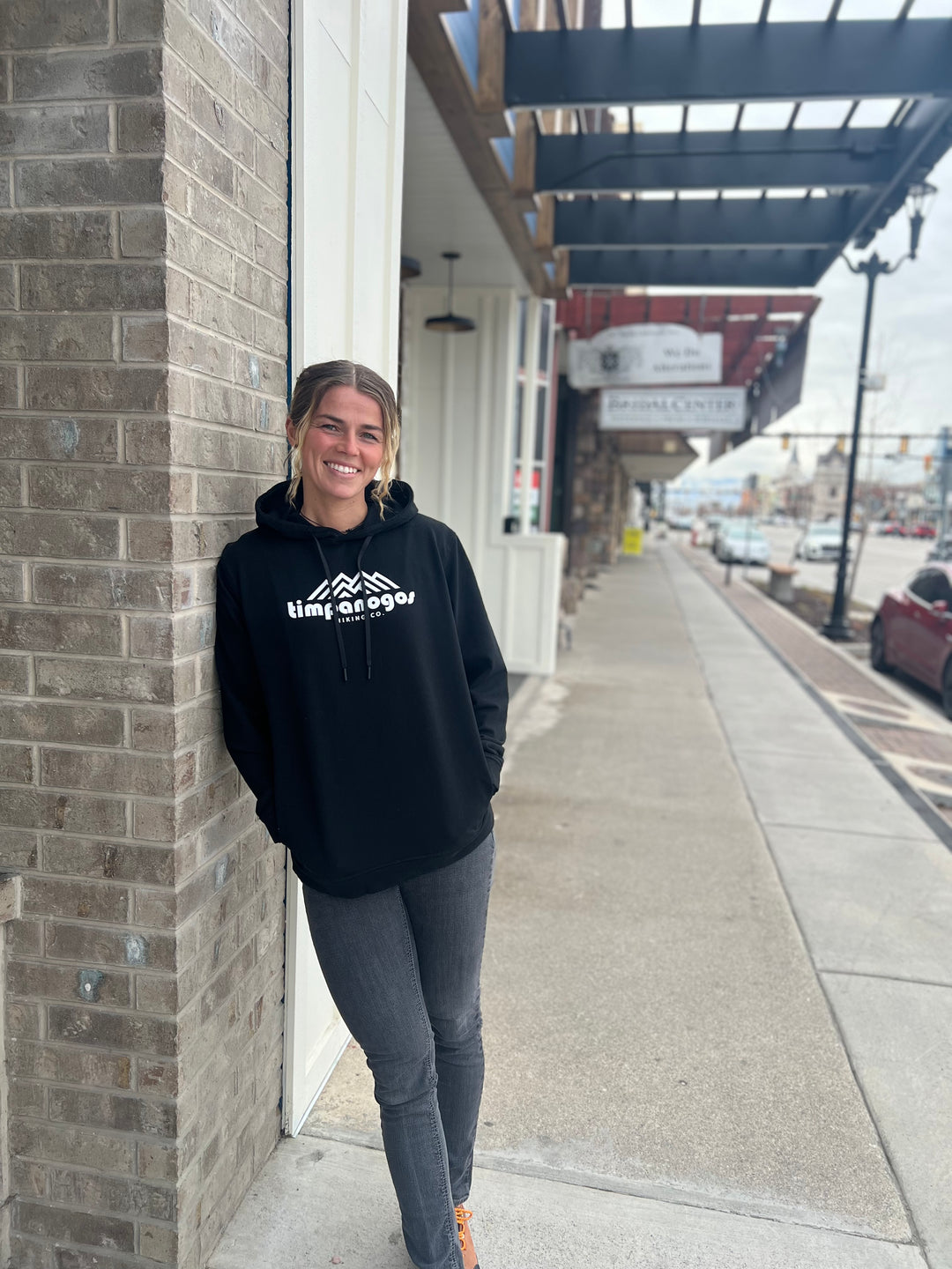 Woman smiling in a black hoodie with the Timpanogos Hiking Co. logo, standing outdoors against a brick wall with a city street backdrop, showcasing the adventurous spirit of Timpanogos Hiking Co.

