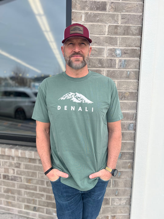 Man wearing a Heather Military Green T-shirt with a white "Denali" mountain graphic, paired with a maroon and white mesh cap, standing outside against a brick wall, representing Timpanogos Hiking Co.'s rugged outdoor spirit.