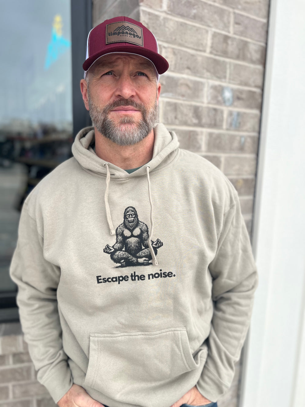 Man in a heather cream hoodie with a meditating Bigfoot and the words Escape the noise., wearing a maroon mesh cap against a brick wall
