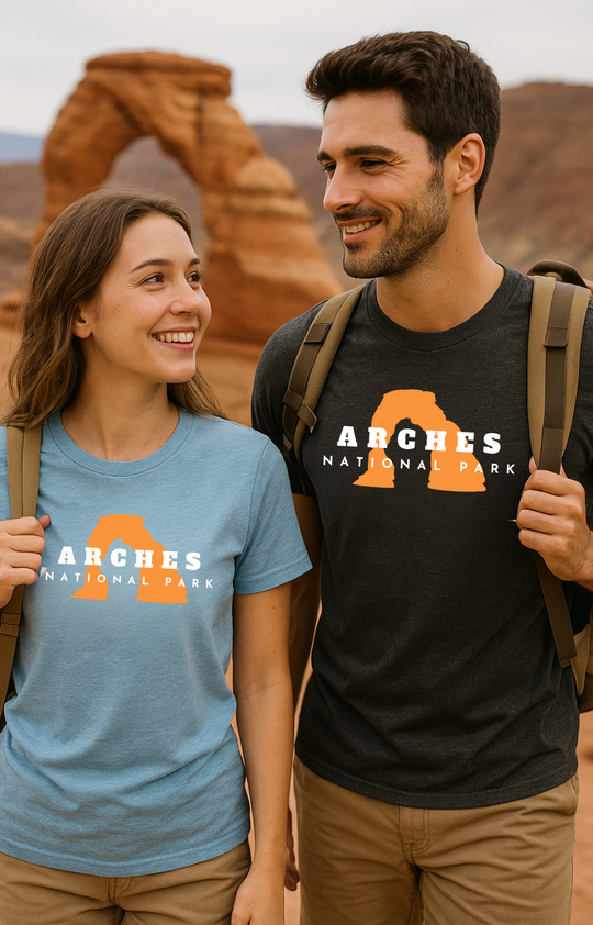Man and woman hiking at Arches National Park, both wearing graphic t-shirts with a bold orange arch design and the words “Arches National Park” in white. The woman wears a light blue shirt; the man wears a charcoal black shirt. Delicate Arch is visible in the background.