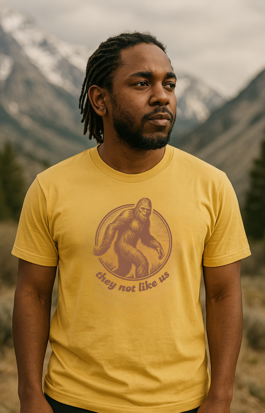 Man standing outdoors in front of snow-capped mountains wearing a mustard yellow t-shirt. The shirt features an illustrated Bigfoot graphic inside a circular frame with the phrase "they not like us" printed below in a vintage-style font. The setting appears to be a rugged mountain valley.